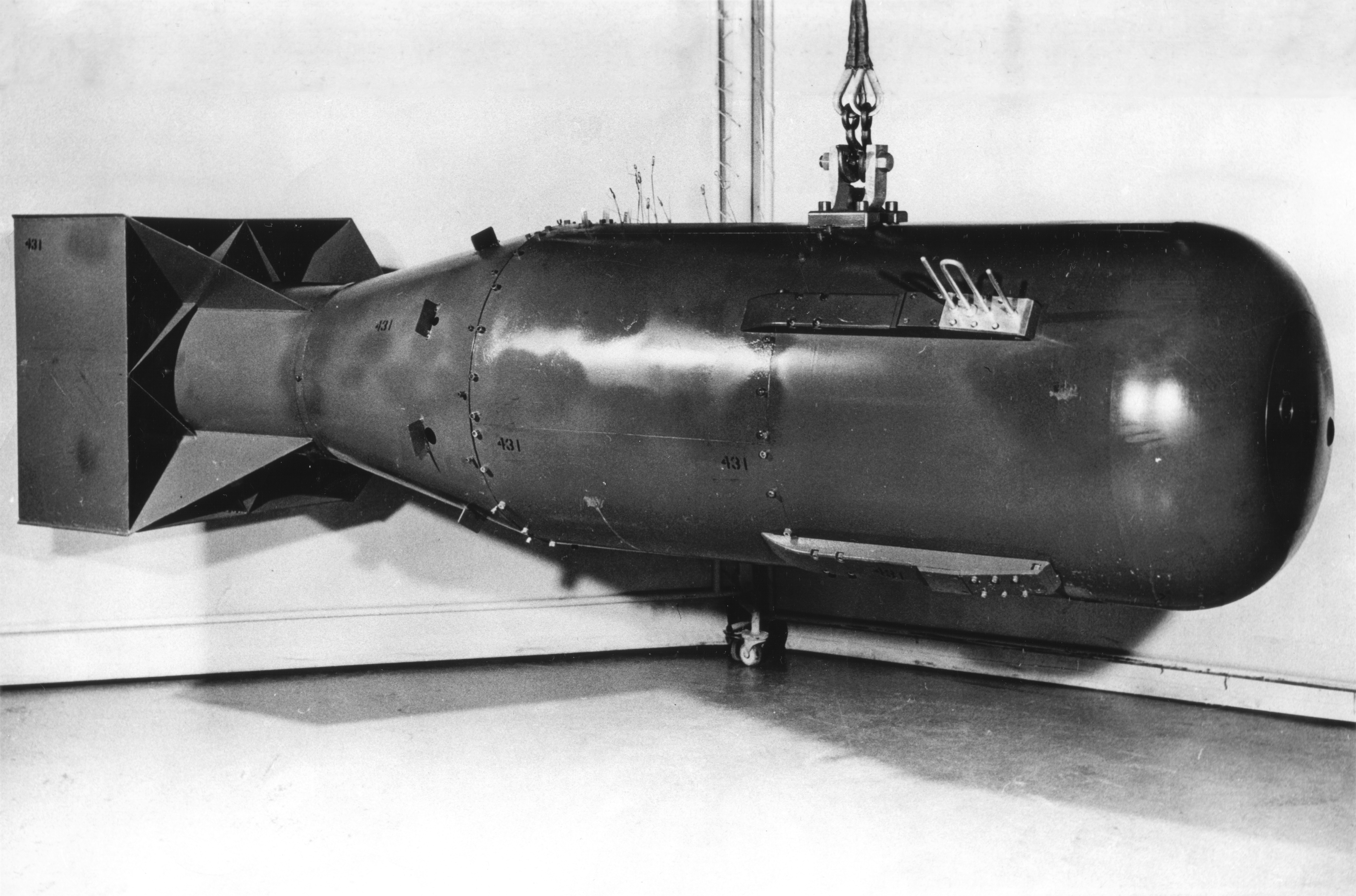 A photo showing the Little Boy bomb on profile. It has the classic bomb shape and appears in dark steel. It is being suspended in the air by a metal cable. 