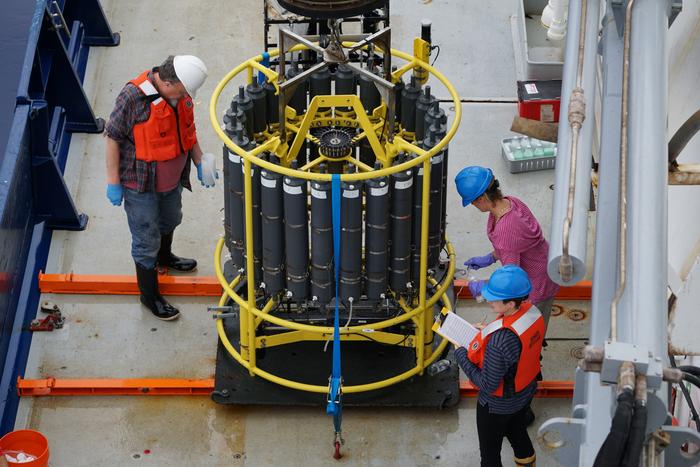 A photo shows a large cylindrical piece of equipment on the deck of a ship. The camera is looking down on the device as three scientists examine it.