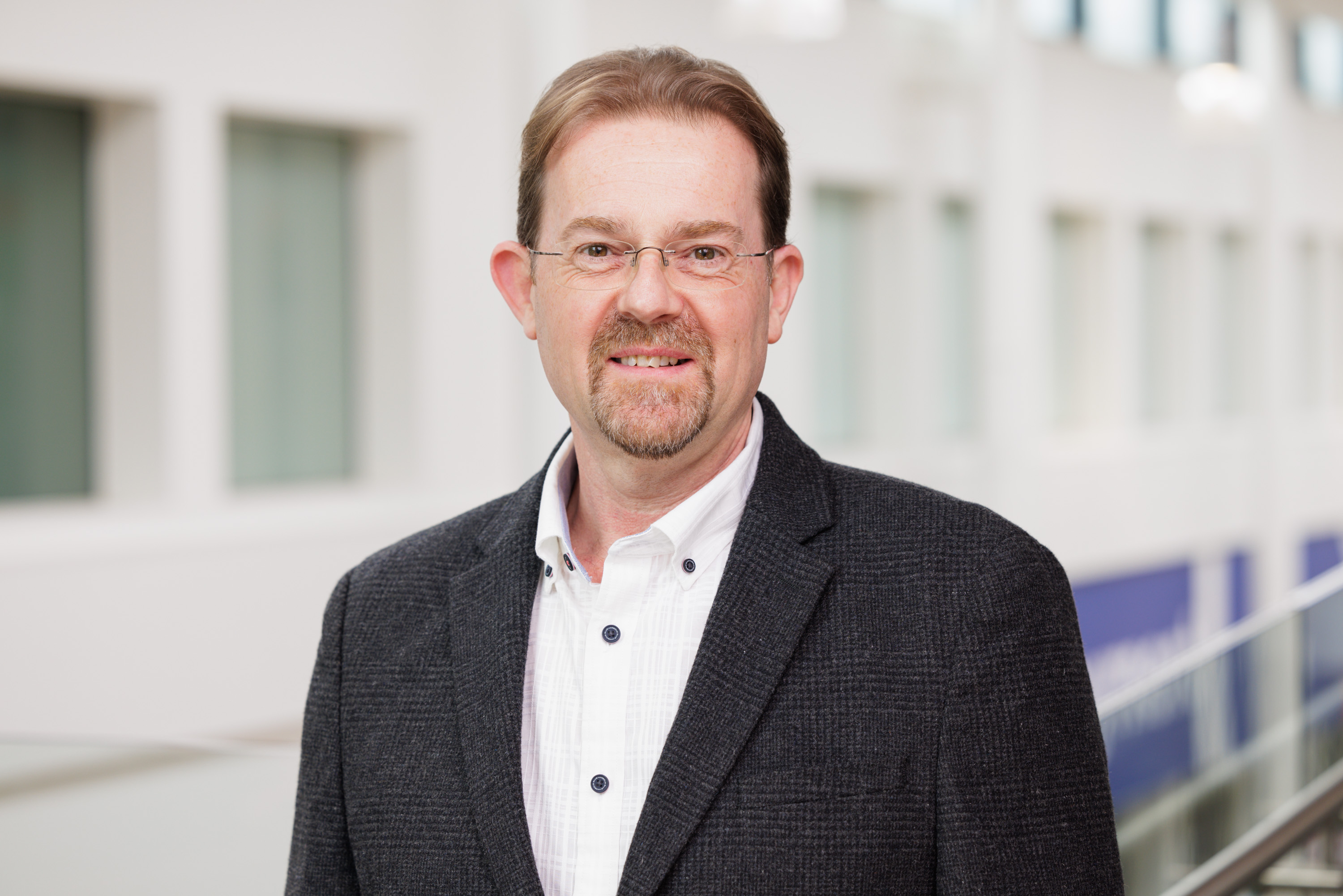 Photo of white man wearing rimless glasses, with pale brown hair and facial hair, wearing a dark tweed jacket and white shirt