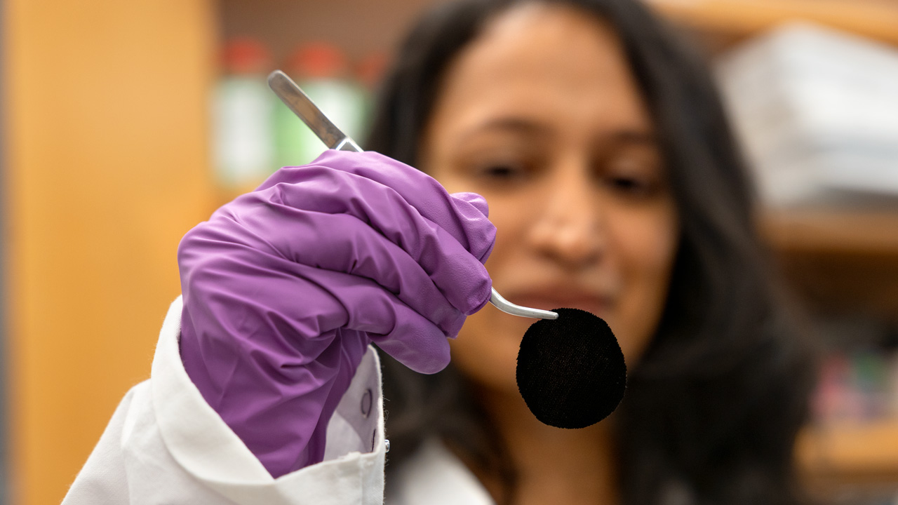 out of focus researcher holding a circle of very dark fabric in a pair of tweezers, while wearing a lab coat and purple gloves out of focus researcher holding a circle of very dark fabric in a pair of tweezers, while wearing a lab coat and purple gloves
