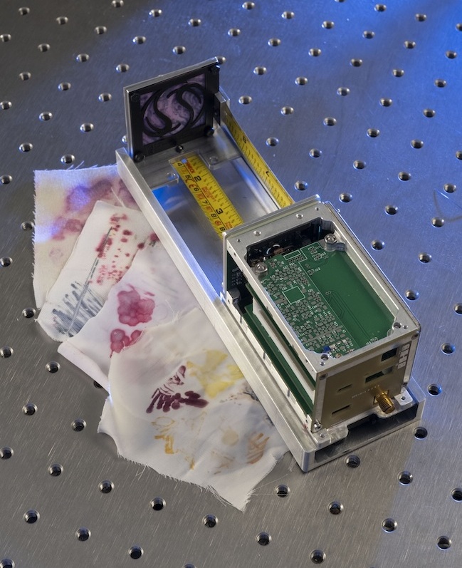 Examples of the fabric painted with bacterial dyes sit beside the Spinning Around SpinnyONE satellite which will carry it into space next year Examples of the fabric painted with bacterial dyes sit beside the Spinning Around SpinnyONE satellite which will carry it into space next year